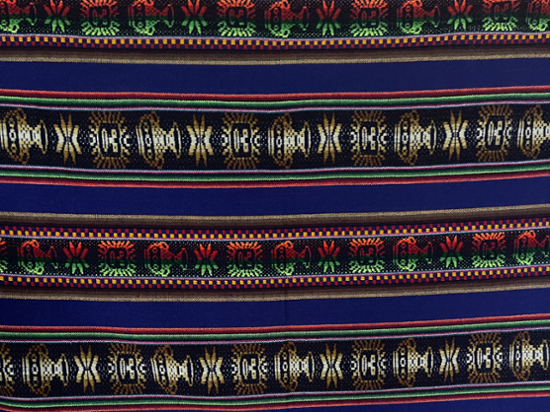 A closeup of richly woven textile with intricate geometric and symbolic patterns in vibrant colors, including deep blue, gold, red, green, and black. The repeating motifs resemble traditional Indigenous or Andean designs, with stylized faces and symmetrical shapes creating a rhythmic visual texture.